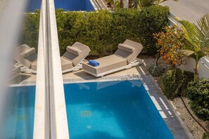 Una piscina al aire libre con sillas de descanso y una mesita, rodeada de follaje y una palmera.