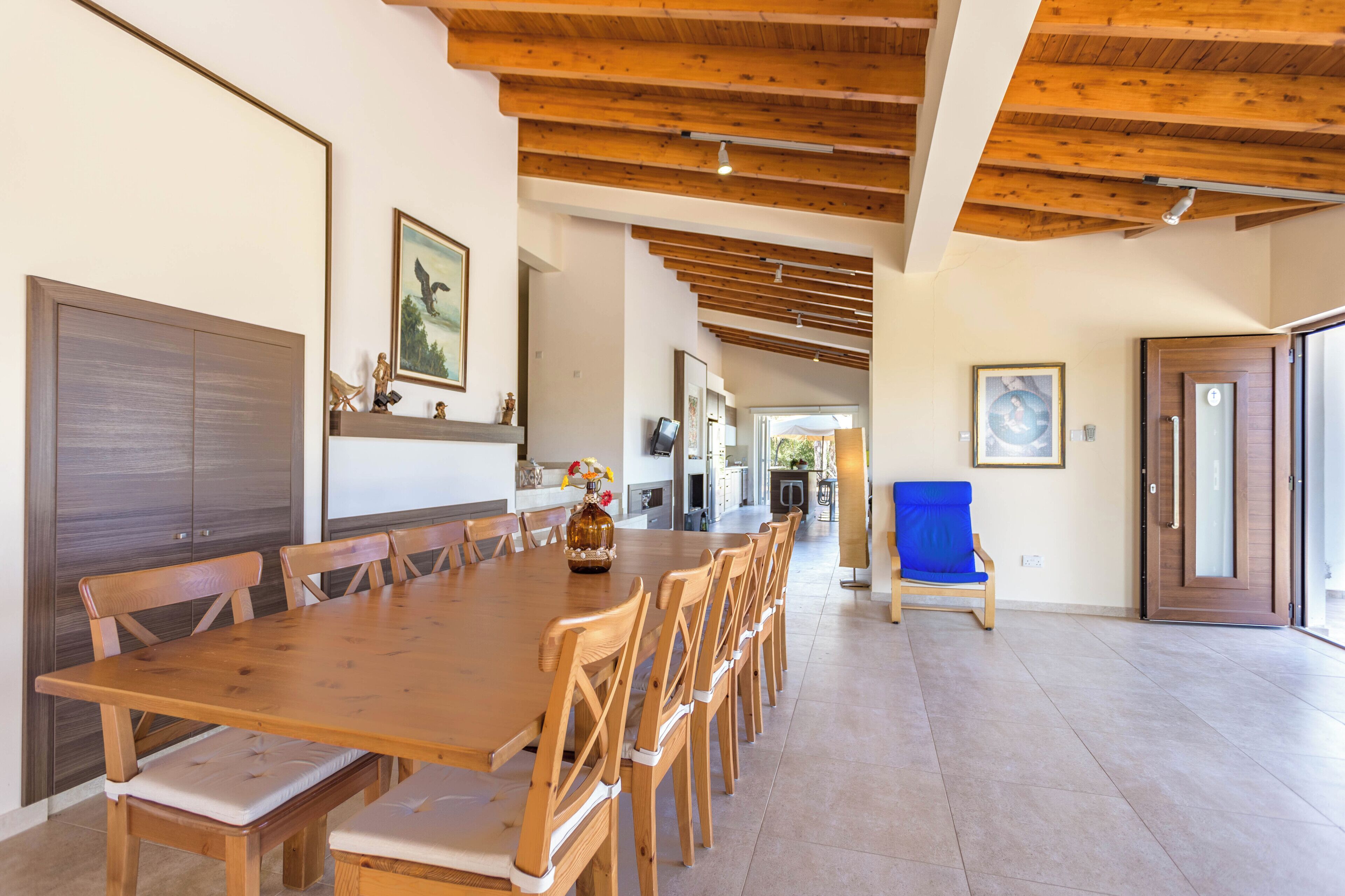 A spacious dining area with a long wooden table and chairs, a blue chair, and a vase with flowers.