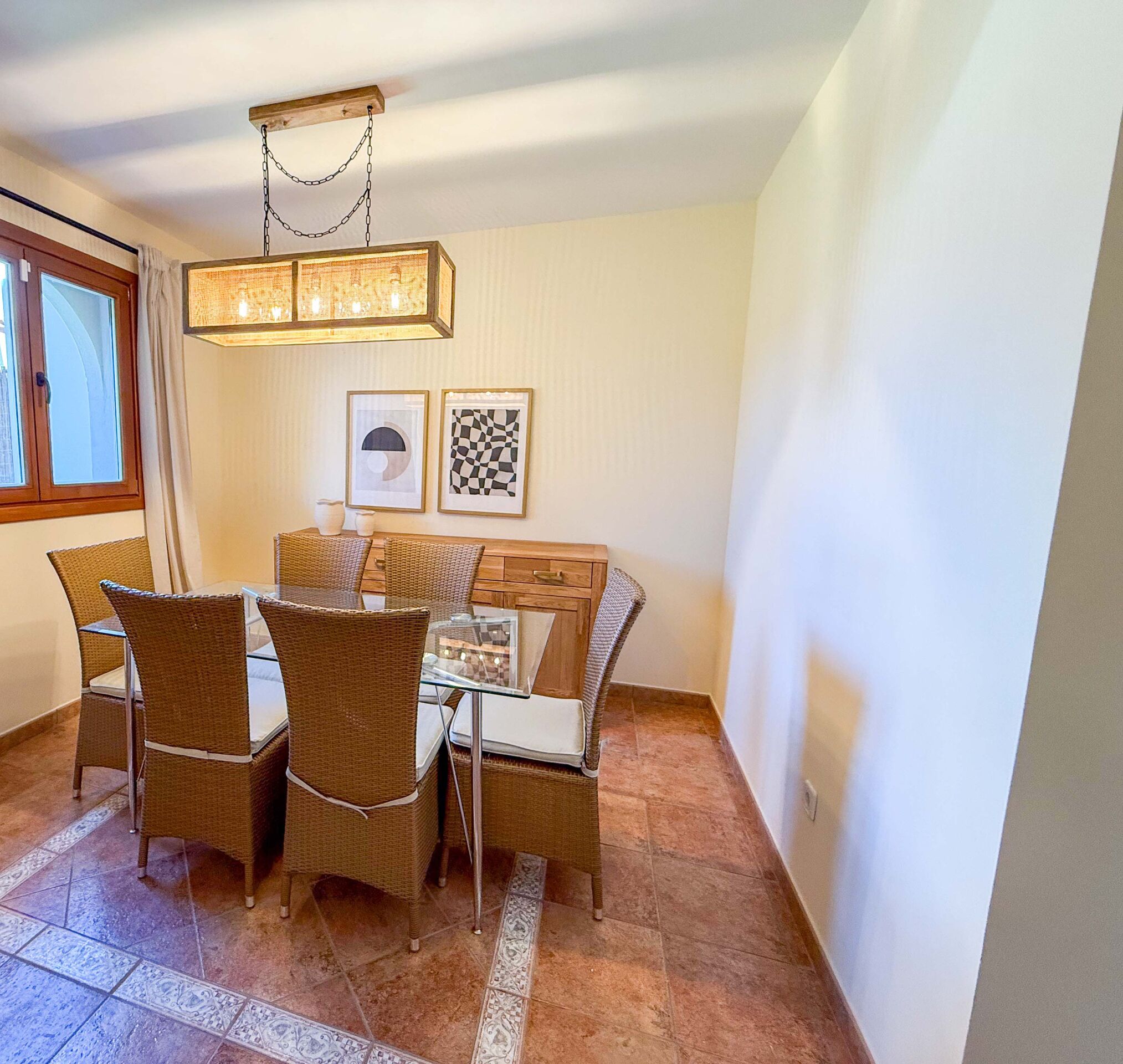 A dining area with wicker furniture, a glass-top table, and two framed artworks on the wall.