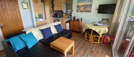 Un espacio habitable compacto con sofá, una pequeña mesa de comedor, kitchenette y televisor montado en la pared.