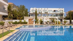 Een modern hotel met een zwembad, ligbedden en een helderblauwe lucht.