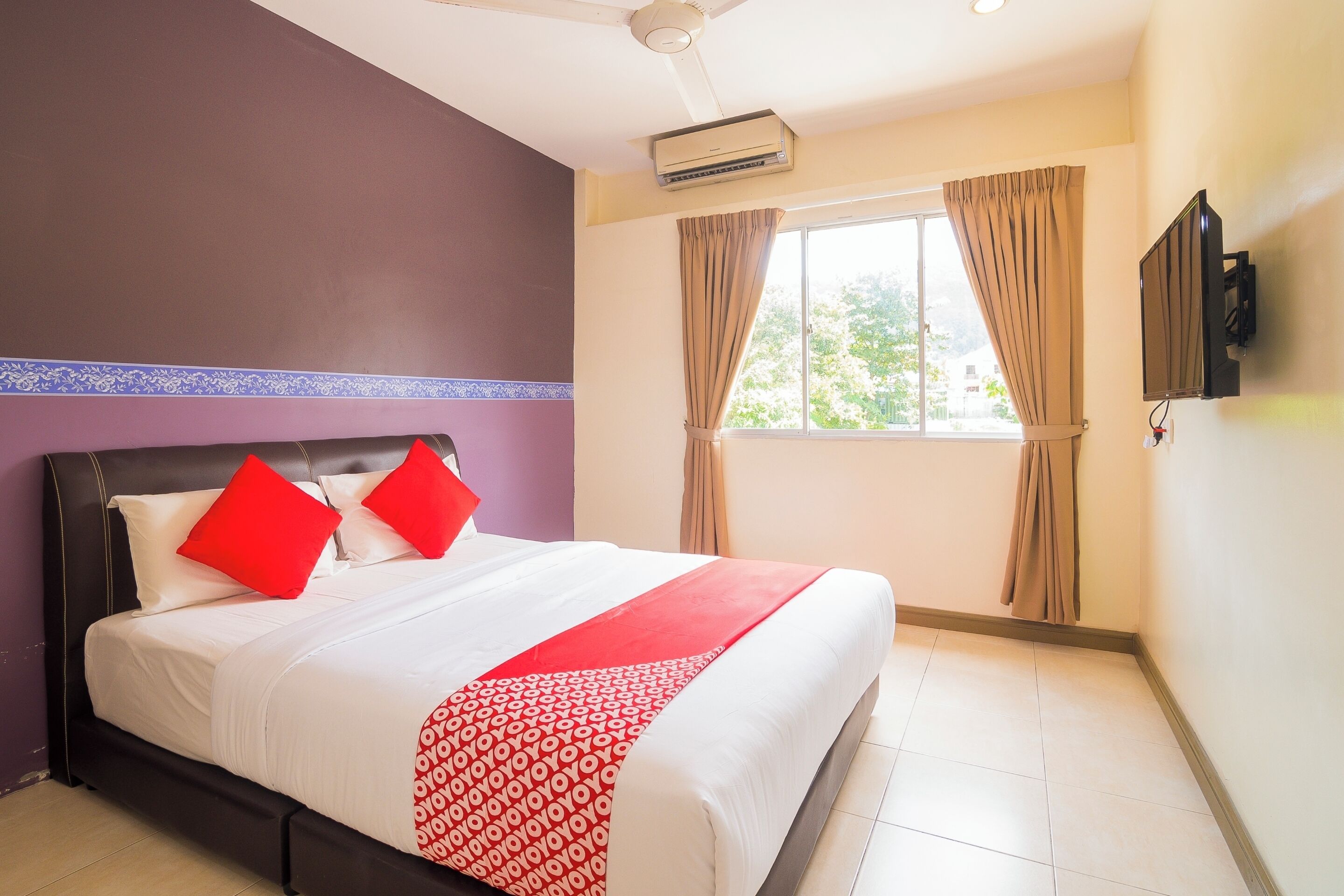 A hotel room with a bed, a ceiling fan, an air conditioning unit, a window with curtains, and a mounted TV.