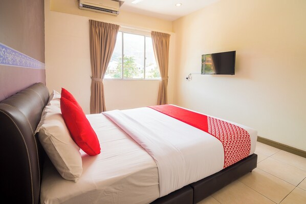 A hotel room with a bed, a flat-screen TV, a window with curtains, and an air conditioning unit.