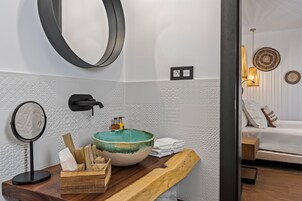 Een badkamer met een grote spiegel, een wastafel met een groene kom, een houten aanrechtblad, en een bed in de aangrenzende kamer.