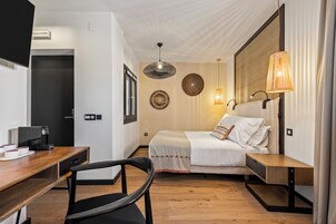 A modern hotel room with a wooden desk, a bed with a white comforter, a wall-mounted lamp, and a ceiling fan.