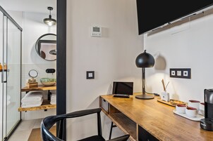 Een moderne kantoorruimte met een houten bureau, een zwarte stoel en een zwarte lamp. Er hangt een spiegel aan de muur, een klein plankje met een schaal, en een koffiezetapparaat staat op het bureau.