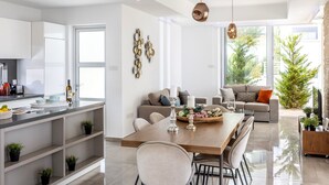 A modern kitchen with white cabinets, a dining area with a wooden table and chairs, and a living room with a sofa and a coffee table. - Milos lifestyle 5 bedroom villa (Protaras)