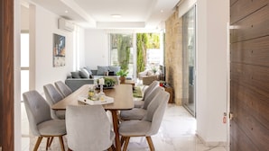 Un comedor moderno con una mesa de madera, sillas tapizadas en gris y un panel de vidrio que separa el espacio del comedor de una sala de estar con un sofá y plantas.