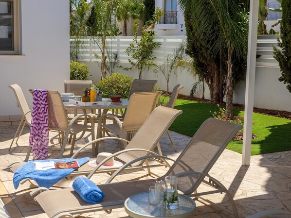 An outdoor patio with wicker furniture, a table with a drink and fruits, and a small garden area. - 3 bedroom villa with pool (Protaras)