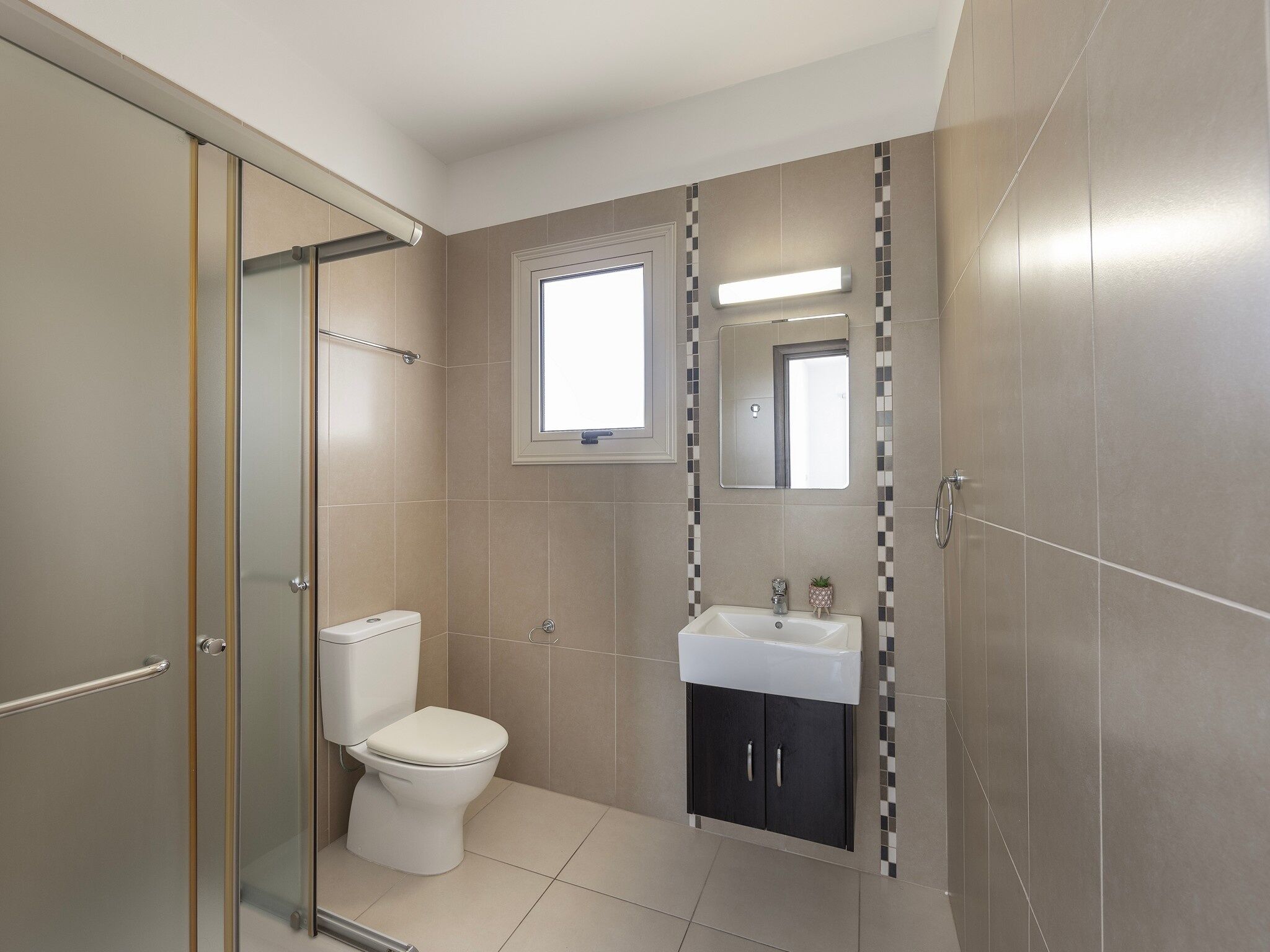 A modern bathroom with a glass shower enclosure, a toilet, a sink with a mirror above it, and a window providing natural light.