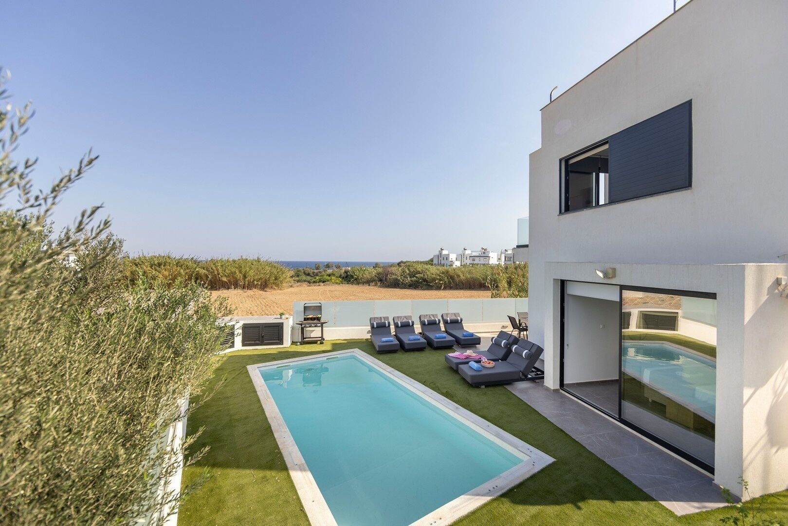 A modern outdoor pool area with a view of the sea, a barbecue, and lounge chairs.