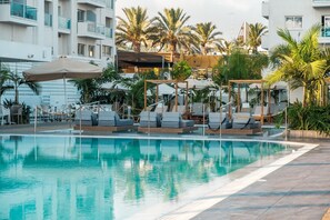 Ein Pool mit Liegestühlen und Sonnenschirmen, umgeben von Palmen und einem mehrstöckigen Gebäude.