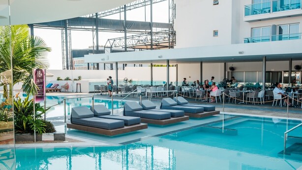 An indoor swimming pool with lounge chairs and tables, surrounded by a covered area with a view of a building. - 1 bedroom apartment in the heart of Protaras (Protaras)