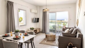 A modern living room with a dining area, a flat-screen TV, a large window with a view of the sea, and a balcony with outdoor furniture. - 1 bedroom apartment in the heart of Protaras (Protaras)