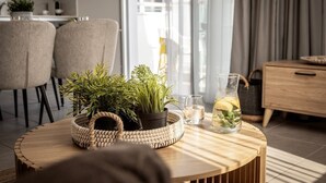 A modern living room with a round wooden coffee table, a woven basket, potted plants, a glass pitcher with a drink, and a dining area with chairs and a table in the background. - 1 bedroom apartment in the heart of Protaras (Protaras)