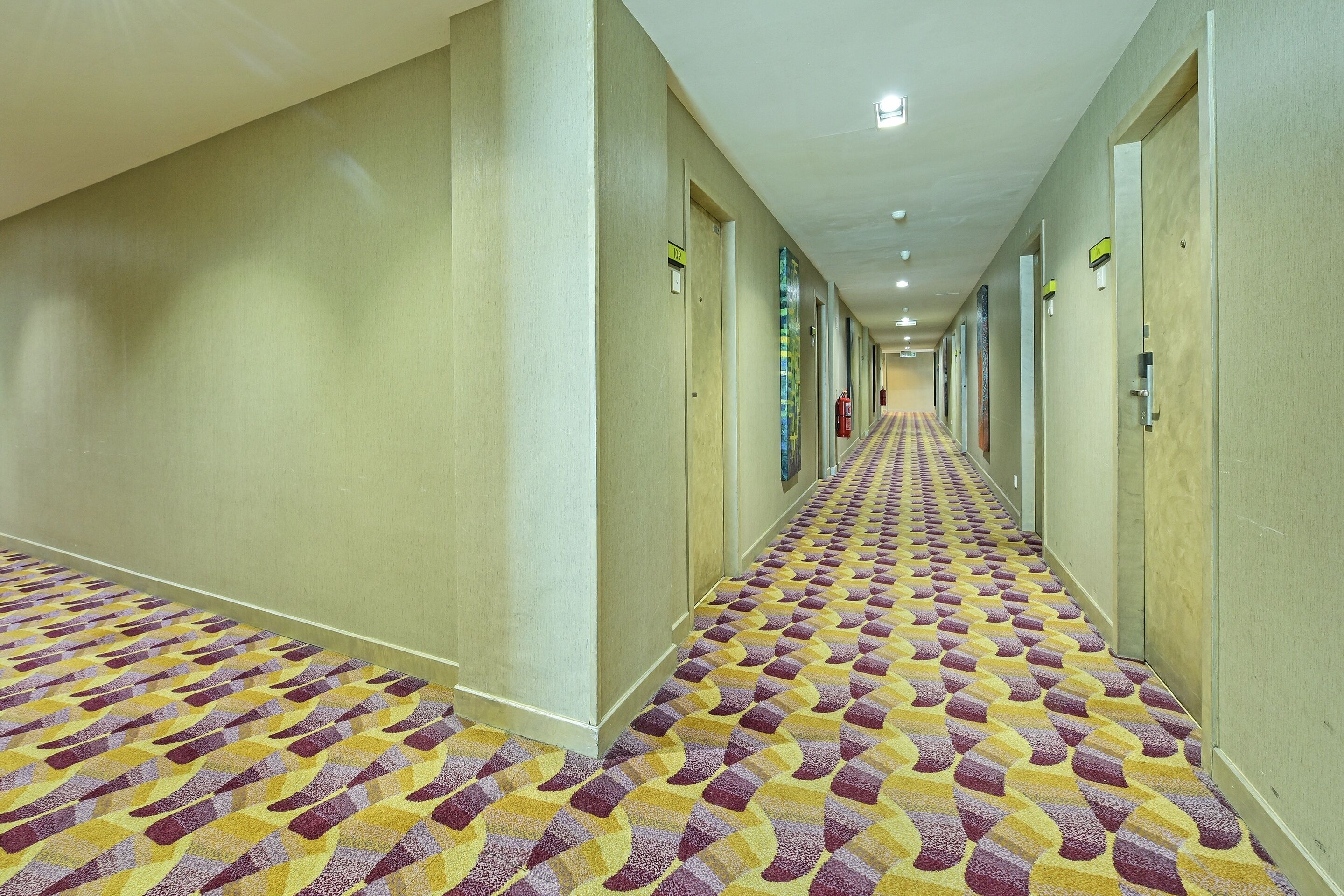 A hotel hallway with patterned carpet, doors on both sides, and a fire extinguisher on the wall.