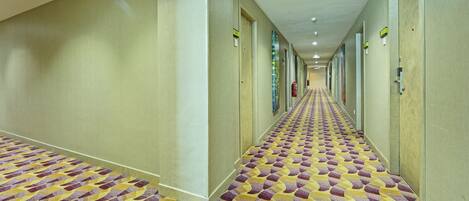 A hotel hallway with patterned carpet, doors on both sides, and a fire extinguisher on the wall.