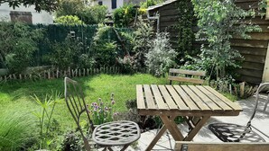 Une table et des chaises en bois, dans un jardin agrémenté d’une clôture en bois, de plantes en pot et de diverses espèces végétales.