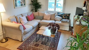 Un salon avec un canapé beige, des coussins à motifs, une table basse en bois et une grande fenêtre donnant sur l’extérieur.