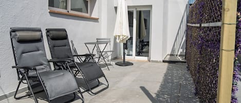 Un área de estar al aire libre con dos sillones de relax y una mesita pequeña, un paraguas blanco y una valla de madera adornada con flores moradas.