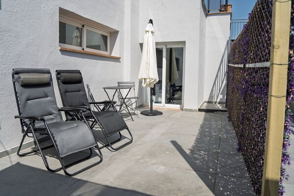 Un área de estar al aire libre con dos sillones de relax y una mesita pequeña, un paraguas blanco y una valla de madera adornada con flores moradas.