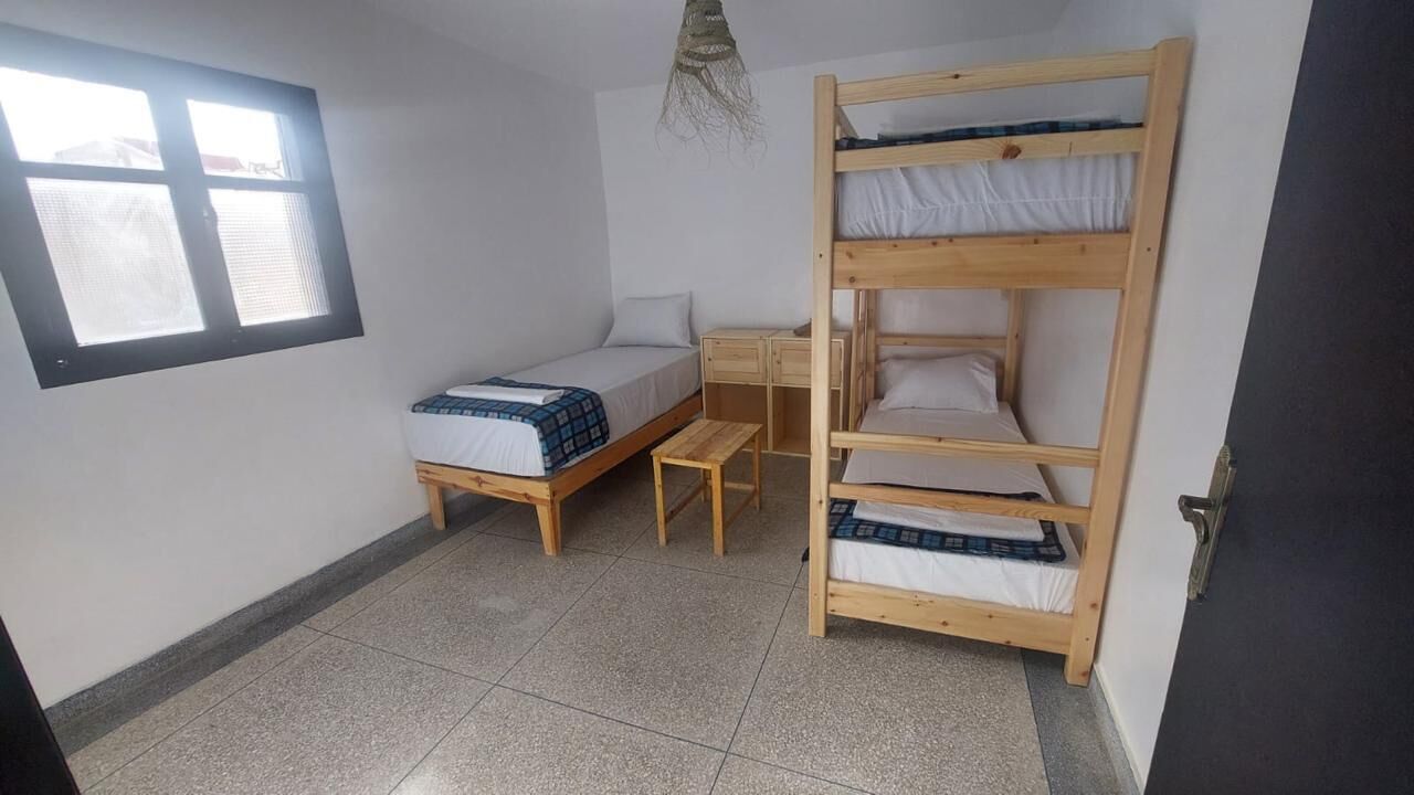 A dormitory room with bunk beds, a single bed, a small wooden table, and a window.