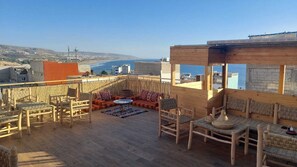 A rooftop terrace with wooden furniture, a small table, and a view of the sea and distant buildings. - HABIBI HOSTEL (Taghazout)
