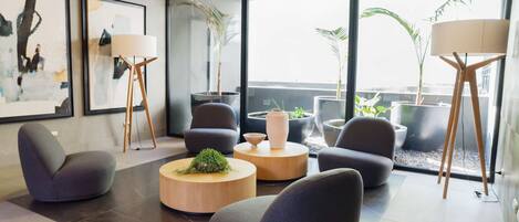 A modern living room with a sofa, armchairs, a coffee table, and floor lamps. There are large windows with a view of the city and plants.