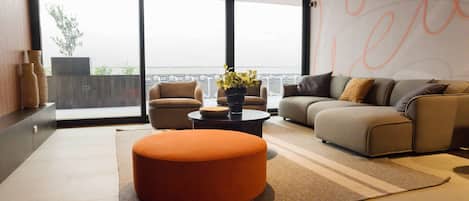 A modern living room with a sofa, ottoman, and coffee table, large windows with a view, and decorative vases.