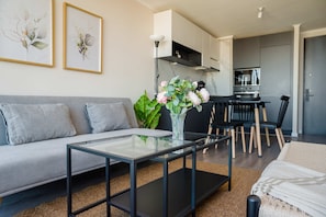 Open-plan living room with a gray sofa, glass coffee tables, and a modern kitchen-dining area accented by botanical artwork and fresh flowers.