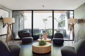 Una sala de estar moderna con un ventanal grande, dos sillones grises, una mesa de centro redonda de madera y dos lámparas de pie.
