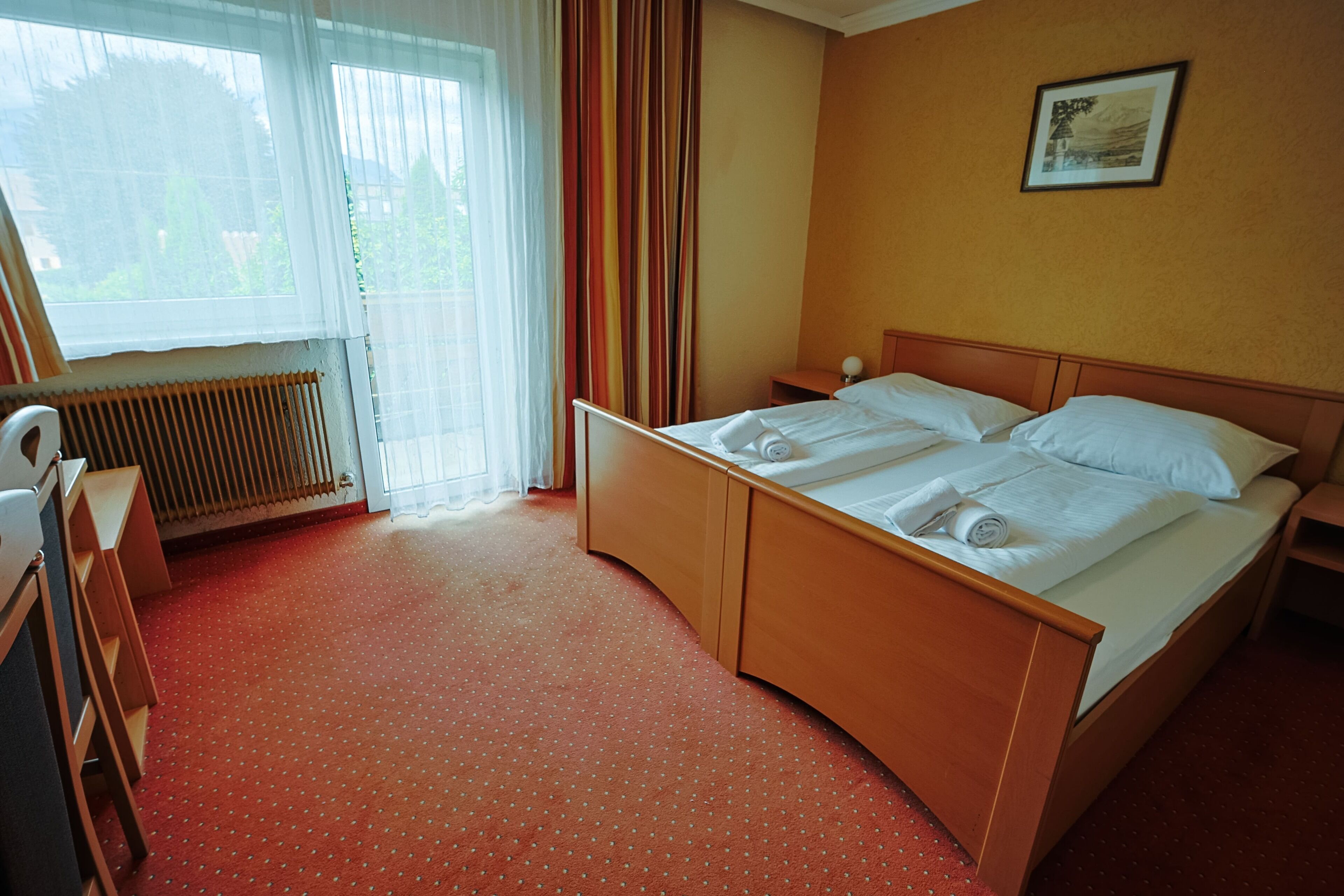 Habitación de hotel con cama de madera, radiador, ventana con cortinas e imagen en el marco en la pared.