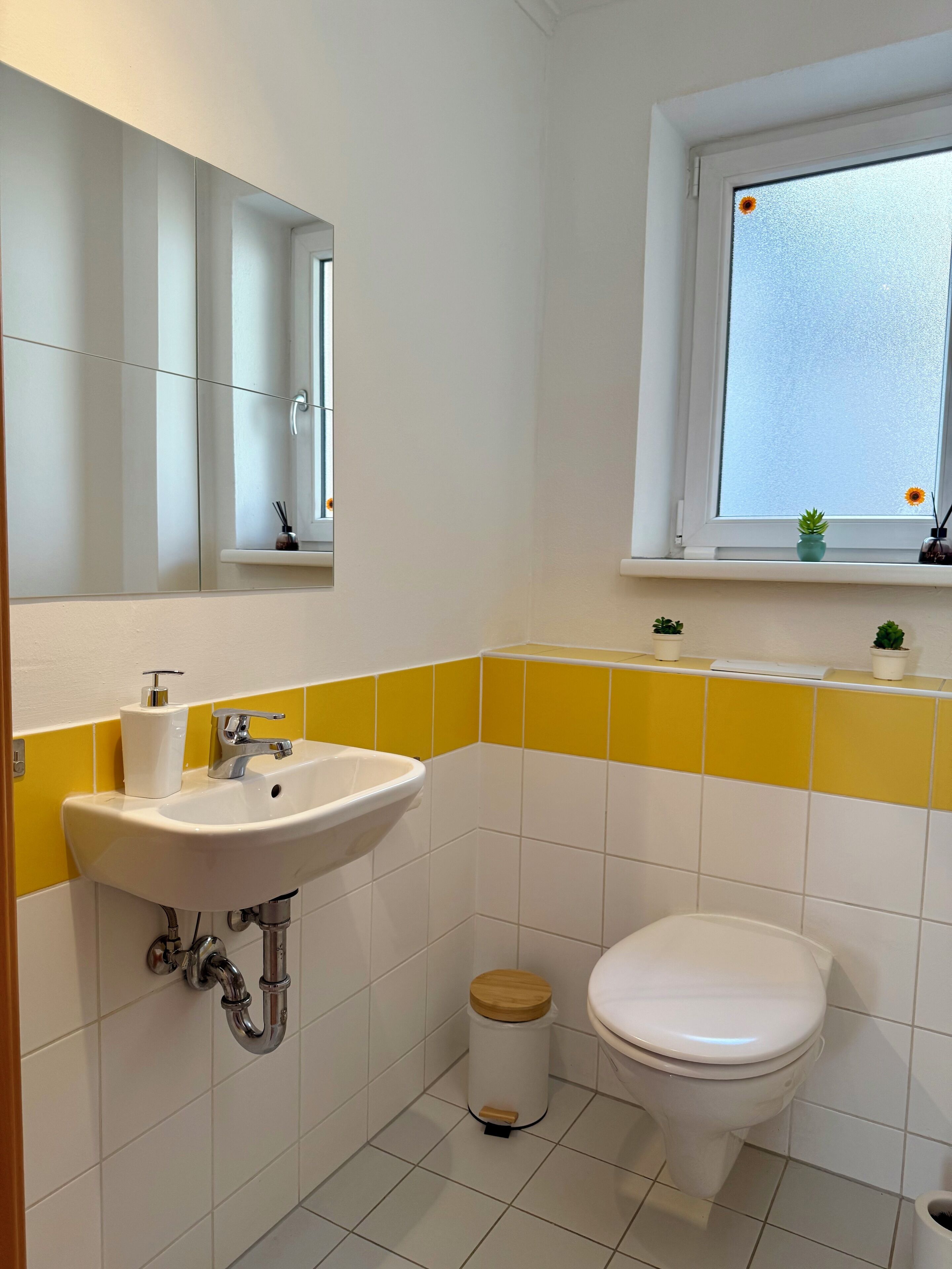 A bathroom with a white sink, yellow and white tiles, a toilet, a window with a view, and a small shelf with a plant.