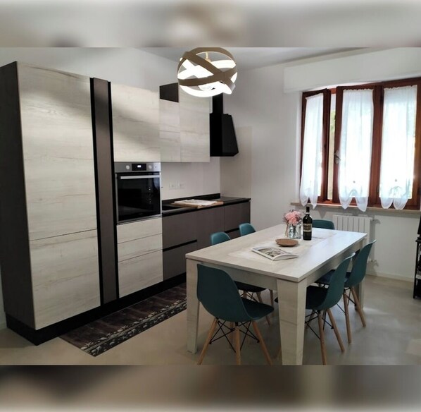A modern kitchen with a built-in oven, a dining area with a white table and teal chairs, and a pendant light above the table. - Modern Trilo residential area live green strategic location sea-center (Senigallia)