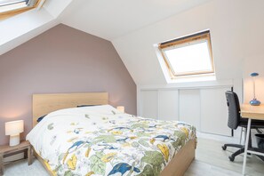 Quarto com teto inclinado, cama com colcha floral, escrivaninha com cadeira e janela.