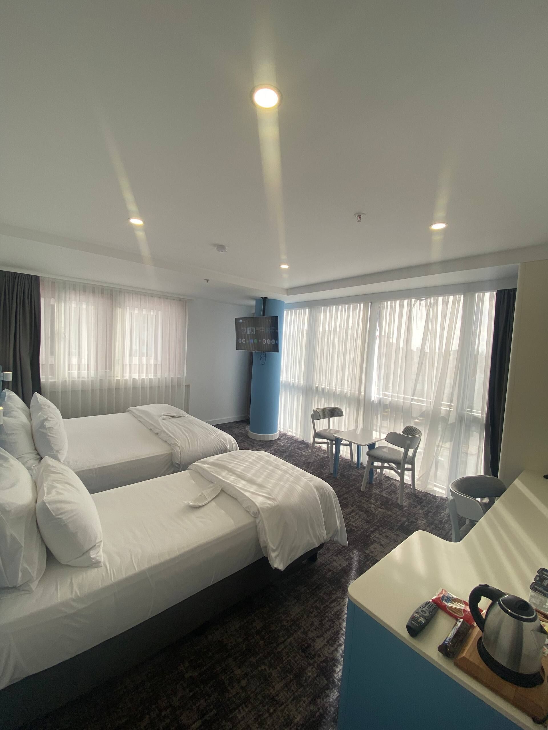 Habitación de hotel con dos camas, televisión, una mesita con sillas y vistas al exterior a través de amplias ventanas.