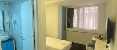 Una habitación de hotel compacta con cama, escritorio con silla, televisión y baño con lavabo y espejo.