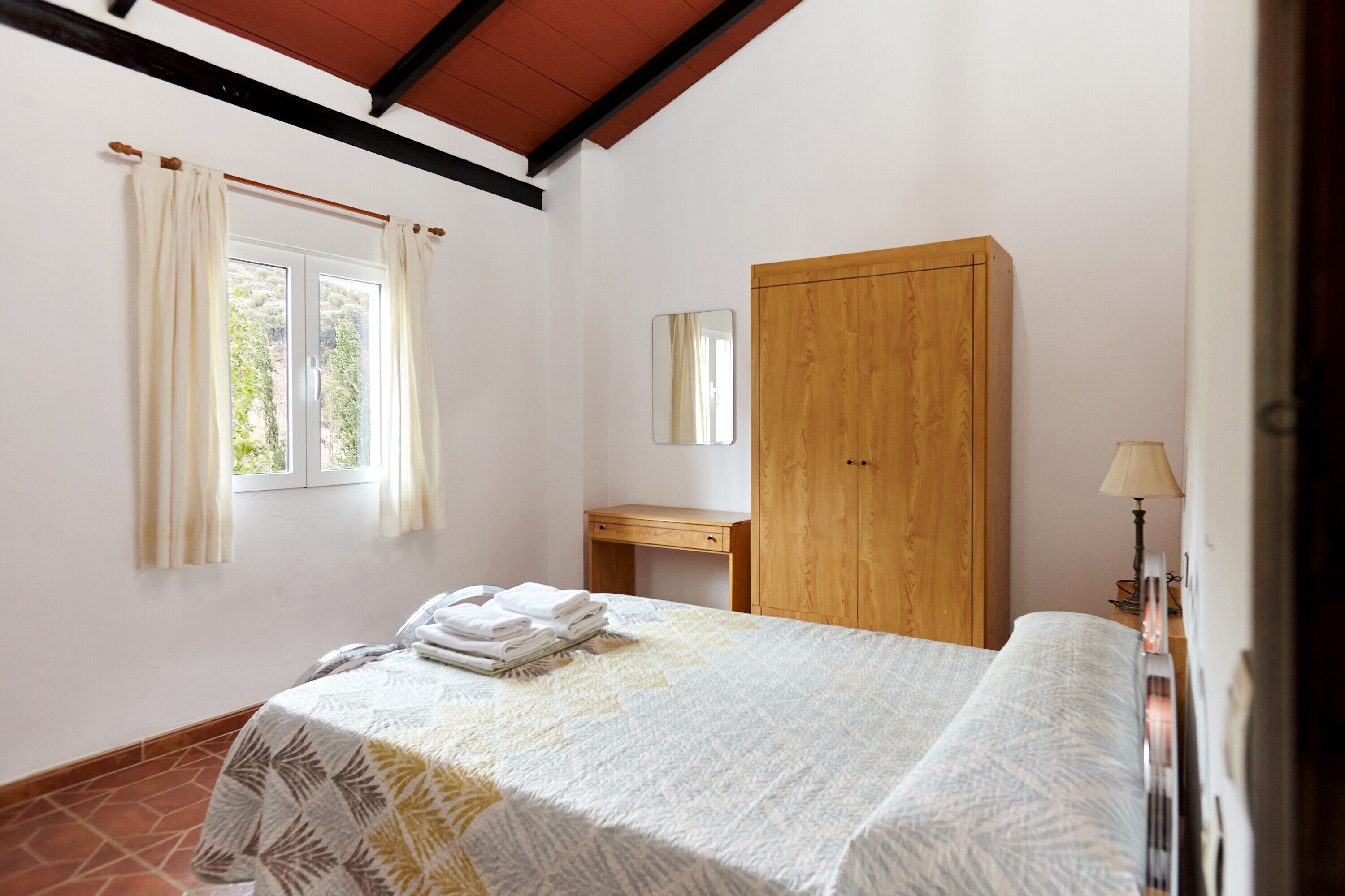 Une chambre à coucher avec un lit, une armoire en bois, un petit bureau et une fenêtre avec des rideaux.