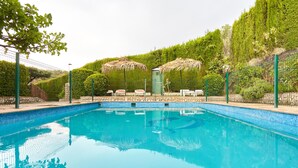A swimming pool with a tiled edge, surrounded by a fence, and shaded by thatched umbrellas.