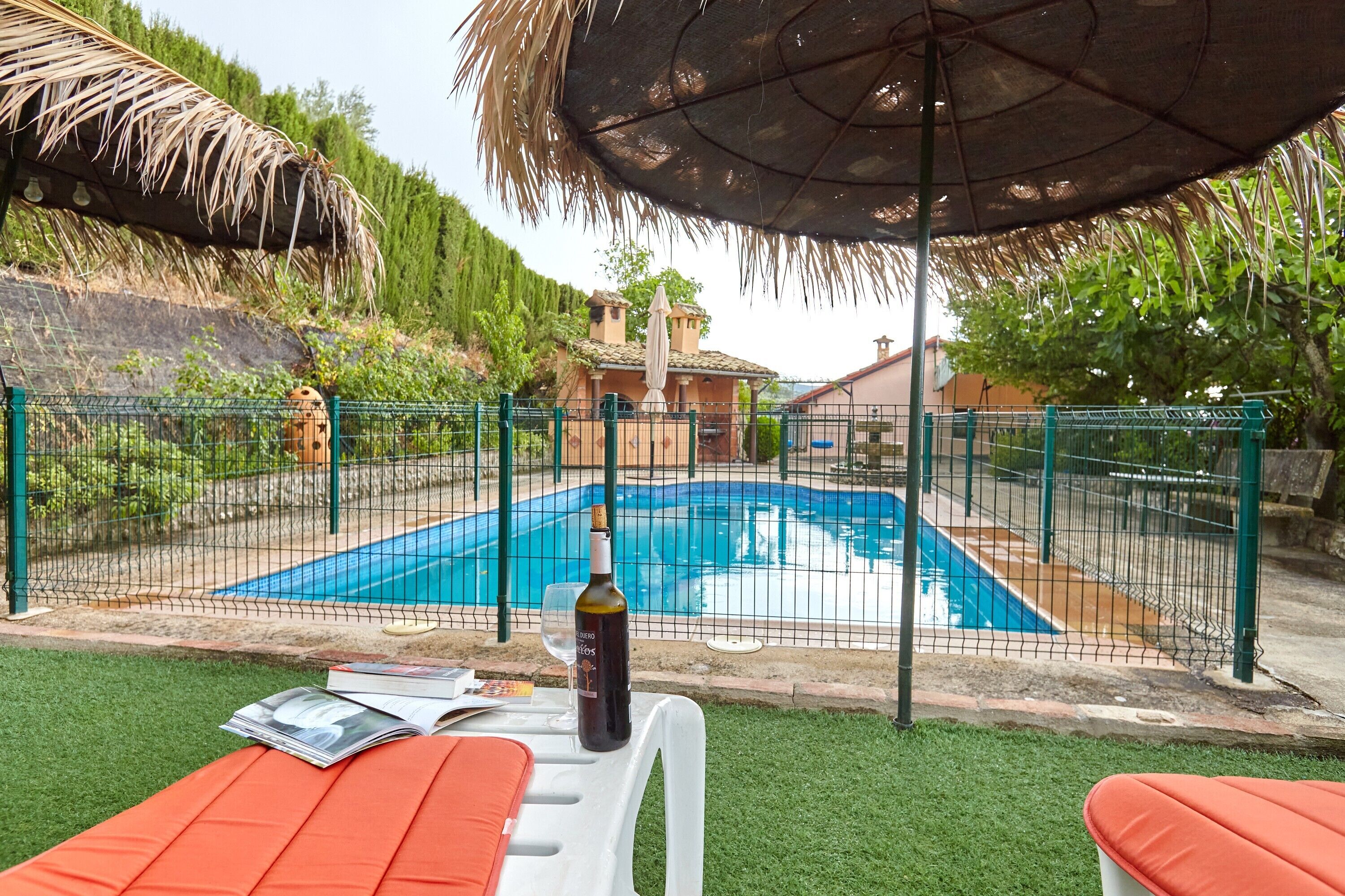 A swimming pool surrounded by a fence, a bottle of wine, and a table with magazines.