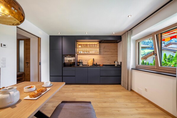 Cocina moderna con armarios oscuros, encimera de madera y ventana con vistas a una casa y vegetación.
