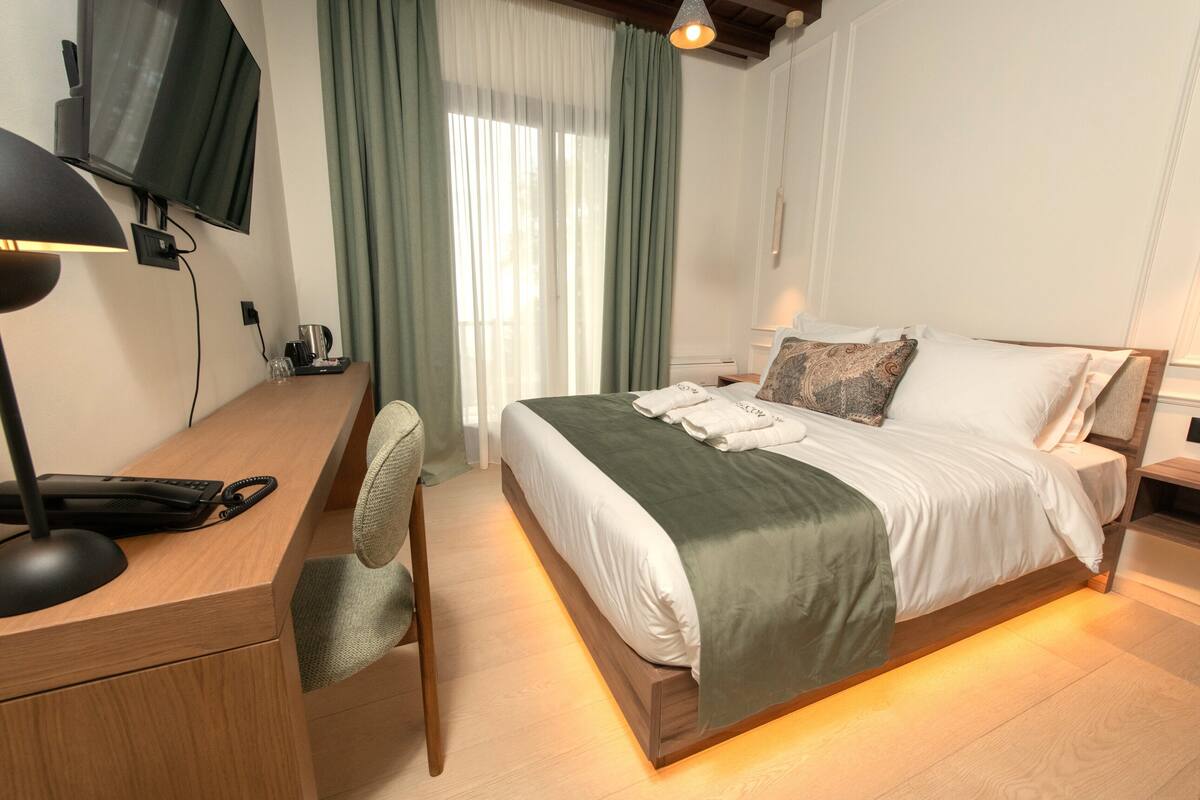 A modern bedroom with a wooden bed, a desk with a chair, a television, and a window with curtains.