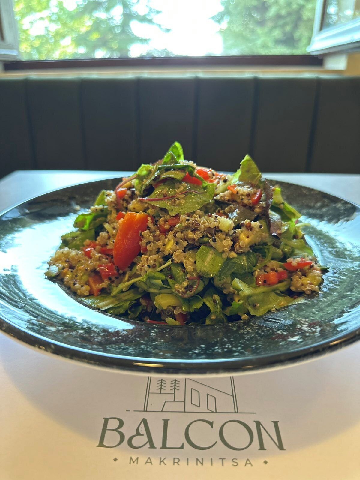 A plate of quinoa salad with vegetables on a table with a logo that reads 'BALCON MAKRINITS'.