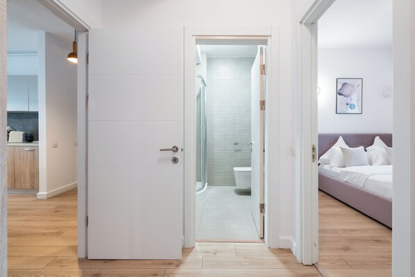 A modern hotel room with a wooden floor, a bed with white bedding, a bathroom with a shower and toilet, and a kitchen area visible in the background. - Uphome Suites (Bucharest)