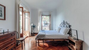 Une chambre avec un plancher en bois, un lit avec une literie blanche, une commode, une chaise, une table de chevet avec une lampe et une fenêtre avec des rideaux.