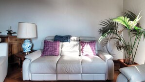 Un salon avec un canapé blanc, un vase bleu et blanc, une lampe et une grande plante.
