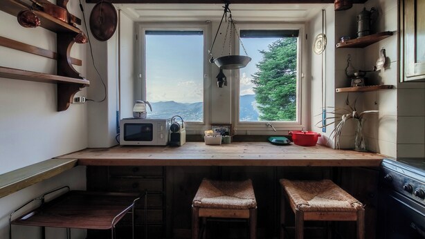 Une cuisine avec des plans de travail en bois, un four à micro-ondes et une fenêtre donnant sur les montagnes. On y trouve deux tabourets en osier et un porte-ustensiles suspendu.
