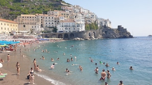 Ein überfüllter Strand mit badenden und sonnenden Personen, ein mehrstöckiges Hotel mit Balkonen sowie eine felsige Küste.