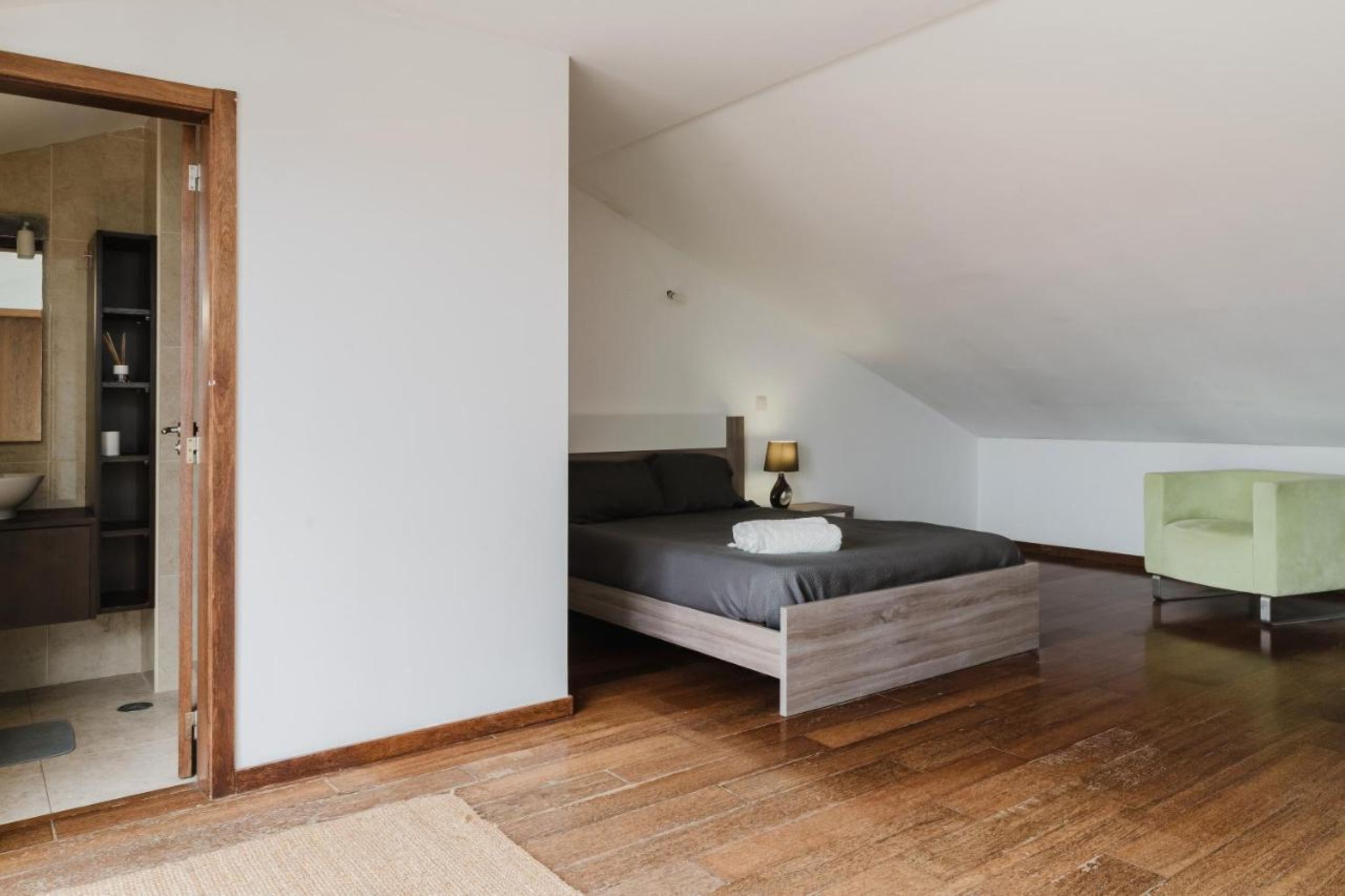 A bedroom with a wooden bed, a bedside table with a lamp, a green armchair, and a view of a bathroom with a wooden cabinet and a towel rack.