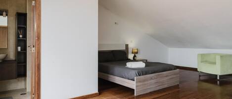 A bedroom with a wooden bed, a bedside table with a lamp, a green armchair, and a view of a bathroom with a wooden cabinet and a towel rack.